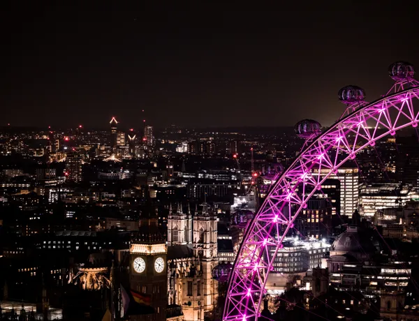 A section of the London Eye with pink lights glowing at night. Landmarks such as the Big Ben and Houses of Parliament can be seen illuminated in the background.