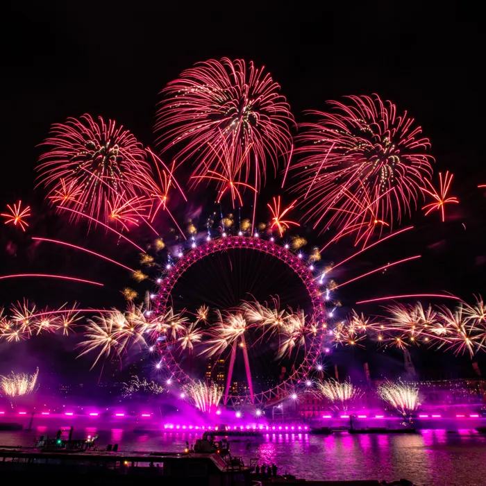 The London Eye lit up in pink at night, with red, yellow and orange fireworks bursting around it during New Year's Eve celebrations.