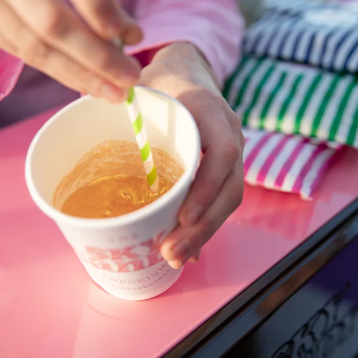 A person leans on a pink table, at the Sky Candy Emporium themed pod at the London Eye, stirring a sparkly gold drink with a green and white striped straw. The table is full of colourful striped paper bags.