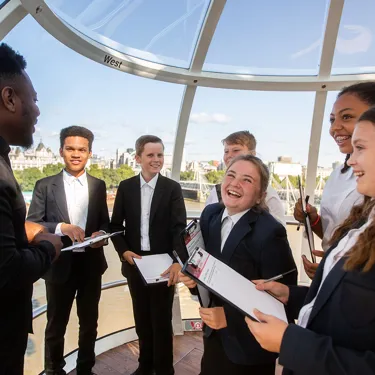 A school group holding clipboards, engaged in a learning activity inside a London Eye Pod, with a view of London landmarks in the background.