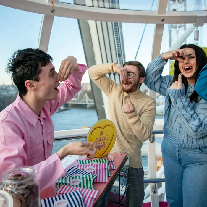 A group of people in The Sky Candy Emporium themed pod on the London Eye. They hold a chocolate button on one hand while pinching their noses with the other.